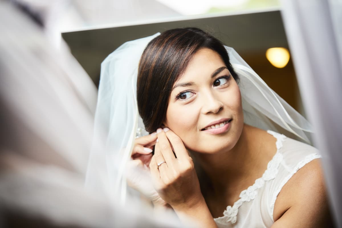 Braut mit Schleier legt vor dem Spiegel ihre Ohrringe an – natürlicher Getting-ready-Moment vor der Hochzeit